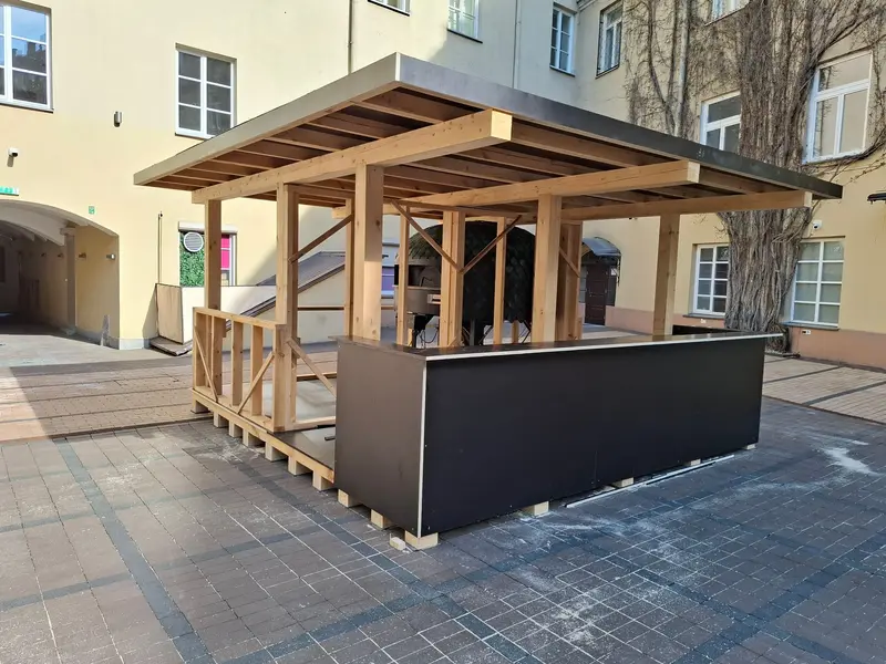 Medinis kiosko karkasas su plokščiu stogu ir tamsiu prekystaliu kieme