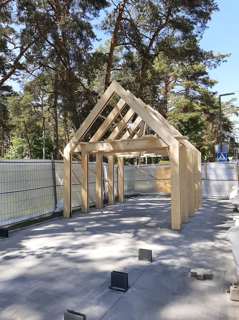 Nedažyti mediniai dvišlaičio paviljono rėmai aptvertoje grįstoje aikštelėje tarp pušų