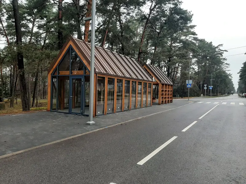 Įstiklintas medinis paviljonas su rudu metaliniu stogu, šalia atviras paviljonas prie kelio