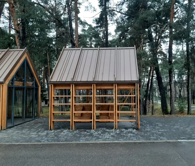 Atviro paviljono šoninė lentynų sienelė ir metalinis stogas, kairėje įstiklintas paviljonas