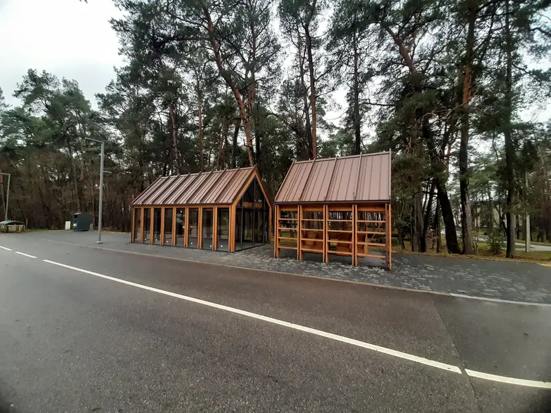 Du mediniai paviljonai su rudais metaliniais stogais pušyne prie asfaltuoto kelio