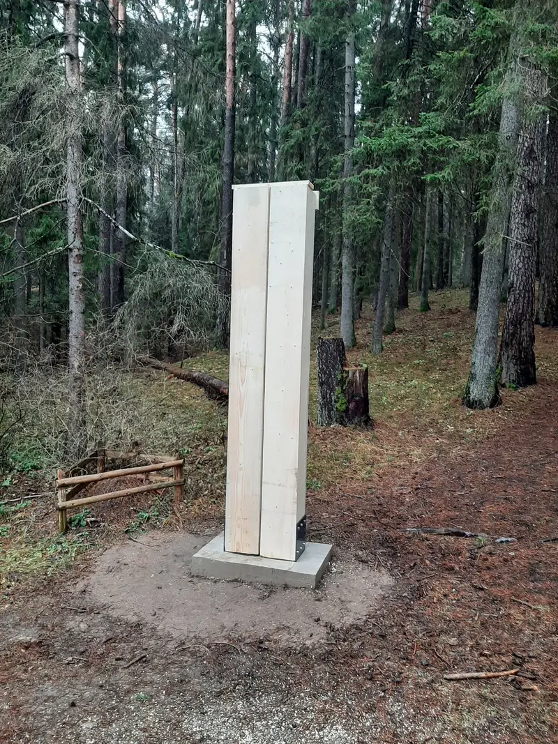Aukštas šviesus medinis stulpas, įtvirtintas metaliniuose laikikliuose ant betoninio pamato miške