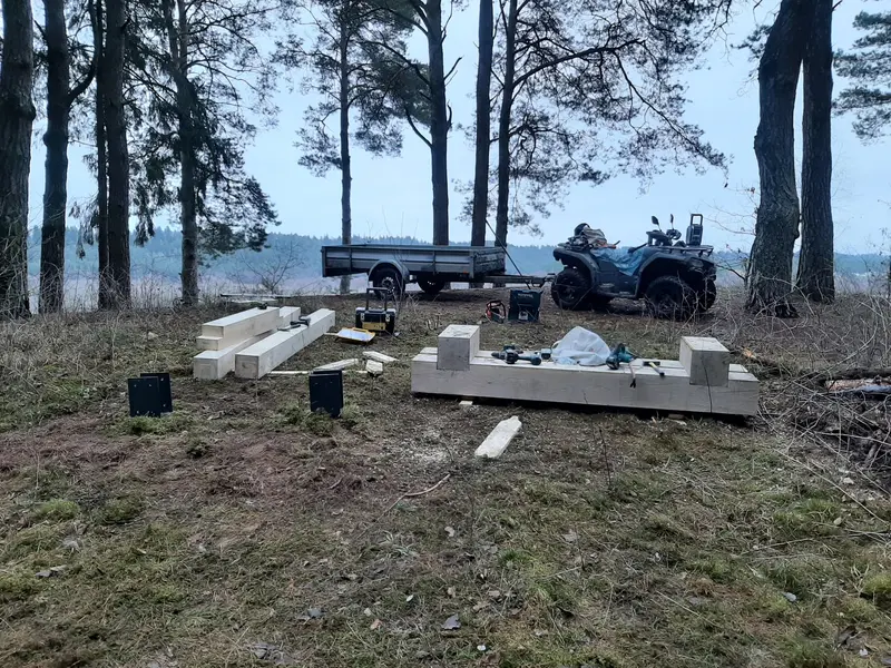 Stambios medienos sijos, metaliniai stulpų laikikliai ir surinkimo metu apverstas suolas pušyne